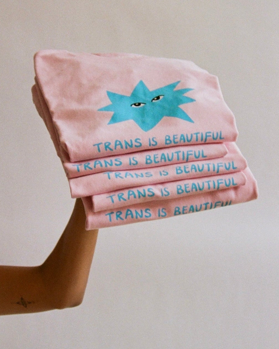 A hand holds a stack of light pink folded T-shirts against a plain light beige background. Each shirt has a blue starburst graphic with small eyes and the text 'TRANS IS BEAUTIFUL' printed in blue.