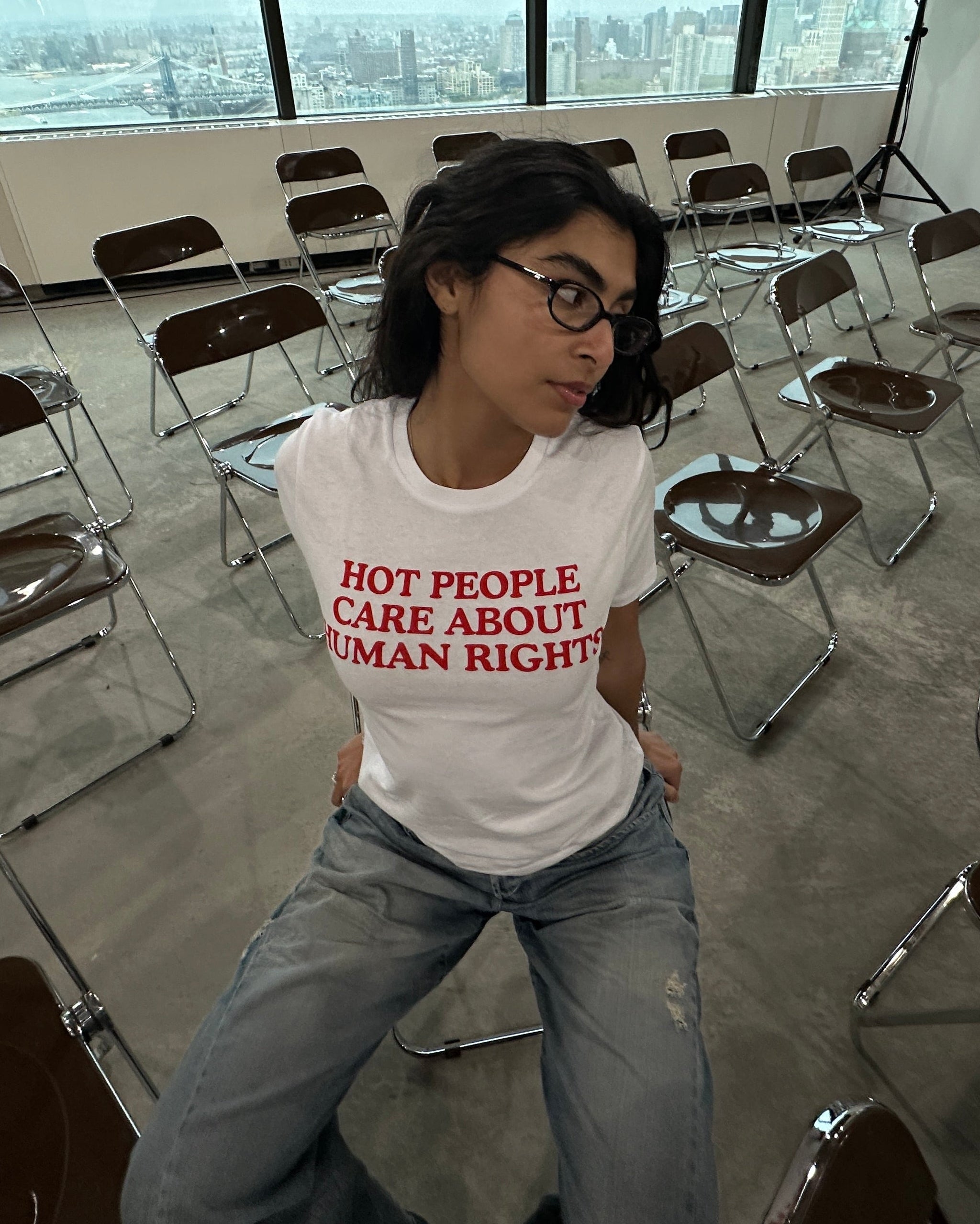 Person wearing a white t-shirt with red text in a room with chairs and large windows.